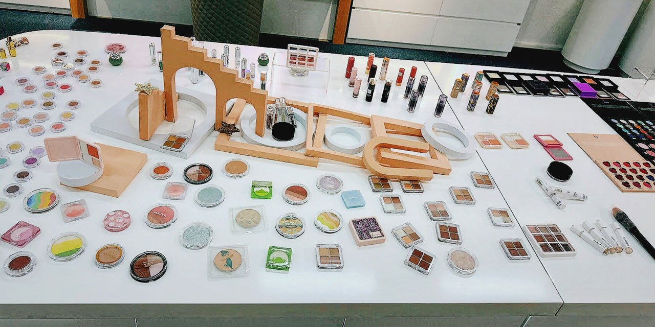 In the showroom, a huge white display table is filled with a variety of colorful blushes, eyeshadows, and powders, presented in a medium shot.