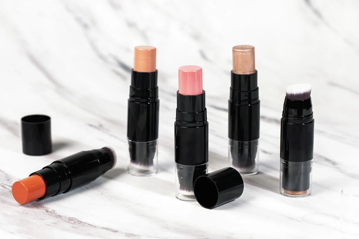Close-up of 4 blush sticks with black tubes and brush tips, in shades like red, peach, and nude, set against a white marble-patterned background