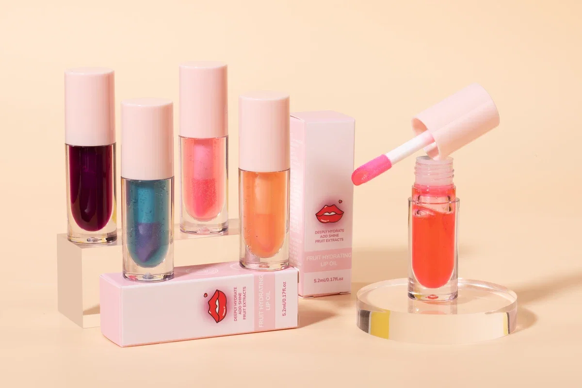 Five Fruit Hydrating Lip Oils in different colors (pink, magenta, green, etc.) standing on acrylic columns. Light yellow background.