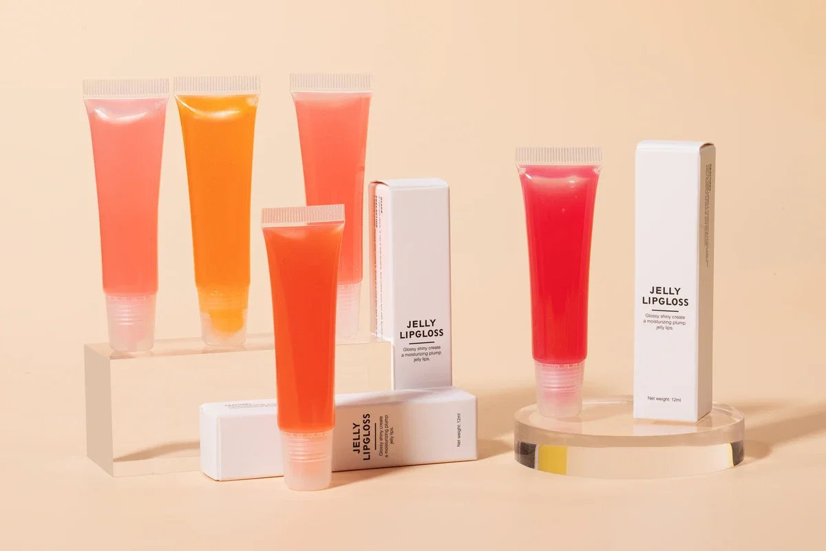 Five Jelly Lipglosses in various colors (peach, blueberry, etc.) along with their white packaging boxes standing on acrylic columns. Light yellow background.